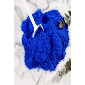 Poudre pigment bleu outremer pour béton ciré décoratif en tas sur pierre, avec spatule blanche et feuilles vertes.