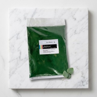 Sachet vert malachite iBéton® pour béton ciré posé sur une plaque marbrée, avec une petite plante à côté.